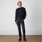 A man wearing a dark navy crewneck sweater, dark wash jeans, a black belt, and brown leather shoes, standing against a light gray wall on a wooden herringbone floor. Oliver Sweeney Sweater available at Sakks Menswear in Southampton.