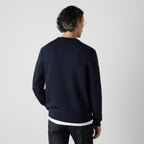 A man viewed from behind wearing a navy blue knit sweater with contrasting white ribbed trim at the collar and hem, paired with dark jeans, against a light gray studio background. Oliver Sweeney Sweater available at Sakks Menswear in Southampton.