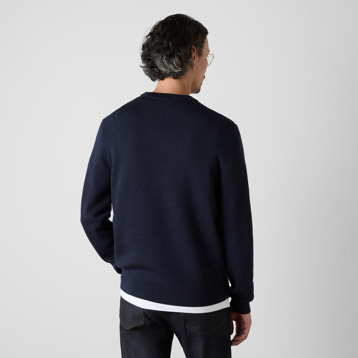 A man viewed from behind wearing a navy blue knit sweater with contrasting white ribbed trim at the collar and hem, paired with dark jeans, against a light gray studio background. Oliver Sweeney Sweater available at Sakks Menswear in Southampton.