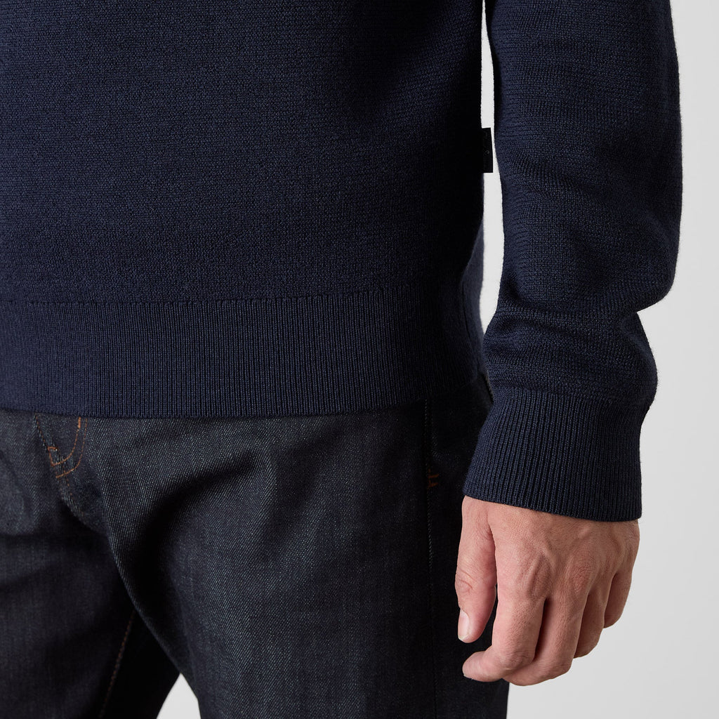 Close-up of a person wearing a dark navy ribbed knit sweater with dark denim jeans, showing the sleeve cuff and hand against a light gray background. Oliver Sweeney Sweater available at Sakks Menswear in Southampton.