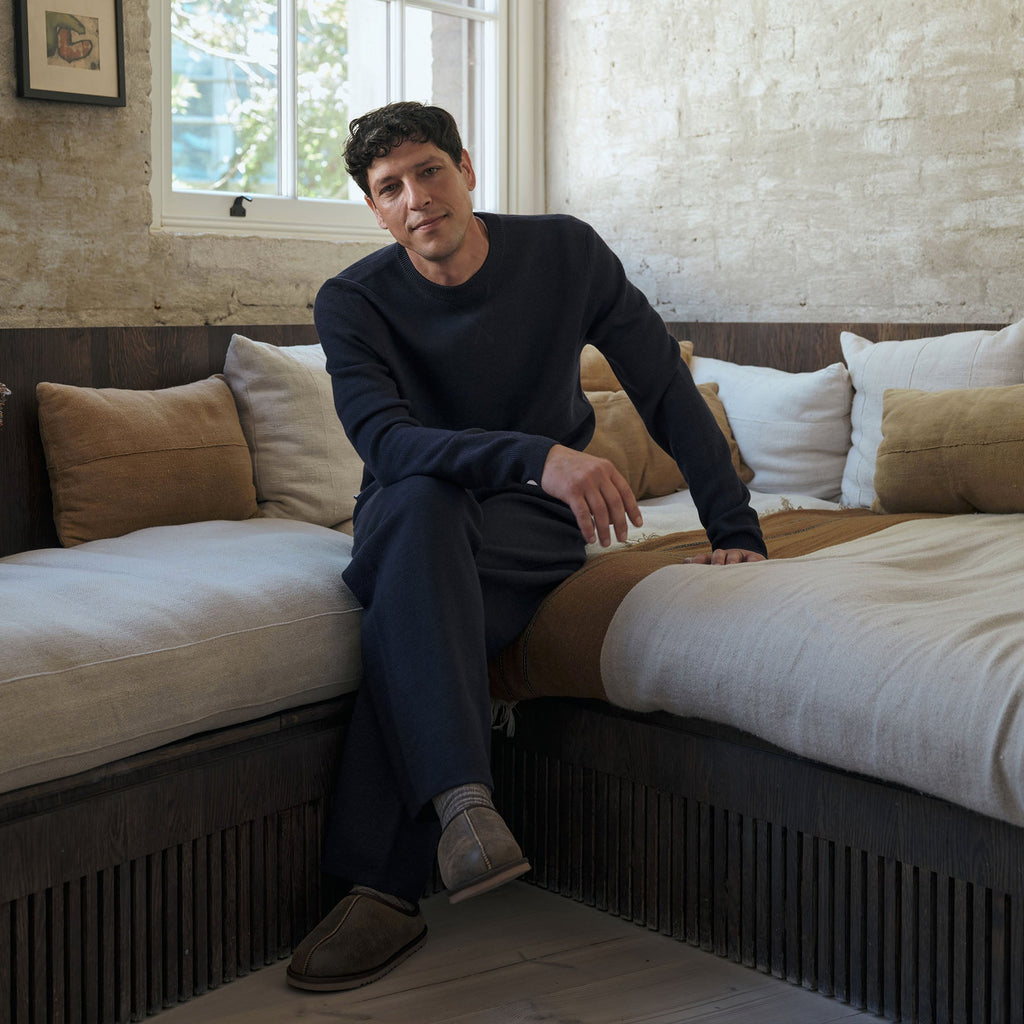 A man wearing a dark navy long-sleeve top and matching navy trousers sits on a daybed in a rustic bedroom setting with neutral-toned pillows and bedding. Oliver Sweeney Sweater available at Sakks Menswear in Southampton.