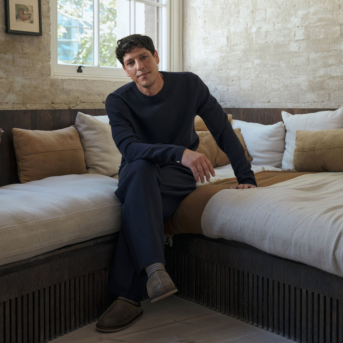 A man wearing a dark navy long-sleeve top and matching navy trousers sits on a daybed in a rustic bedroom setting with neutral-toned pillows and bedding. Oliver Sweeney Sweater available at Sakks Menswear in Southampton.