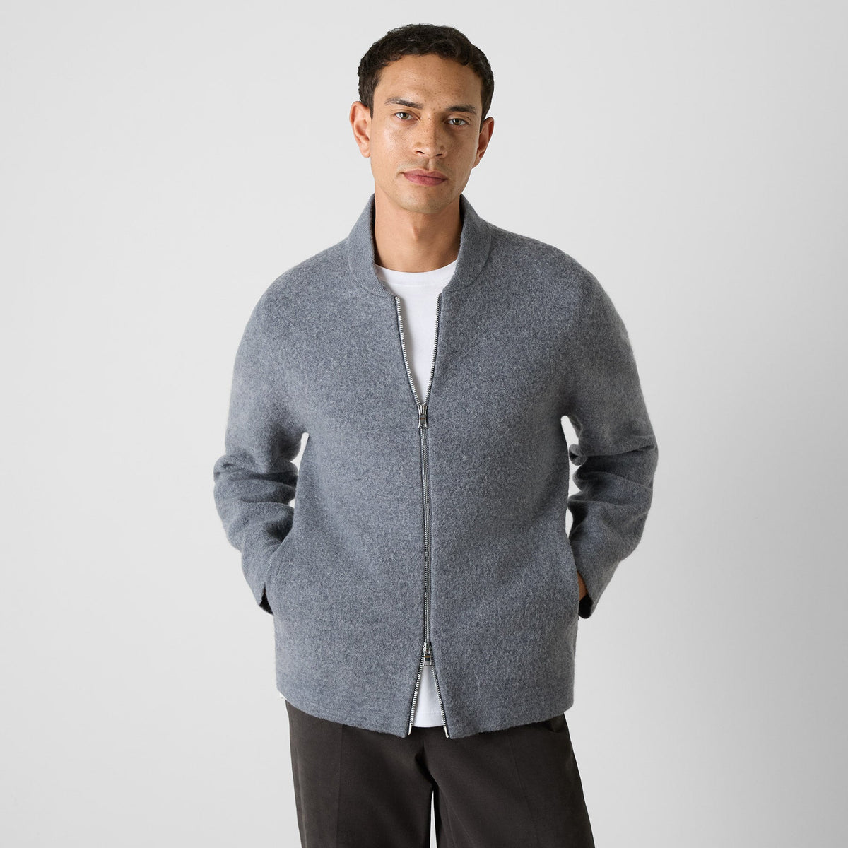 A man wearing a gray zip-up knit jacket with a stand collar over a white t-shirt and dark trousers, standing against a light gray studio background. Oliver Sweeney Cardigan available at Sakks Menswear in Southampton.