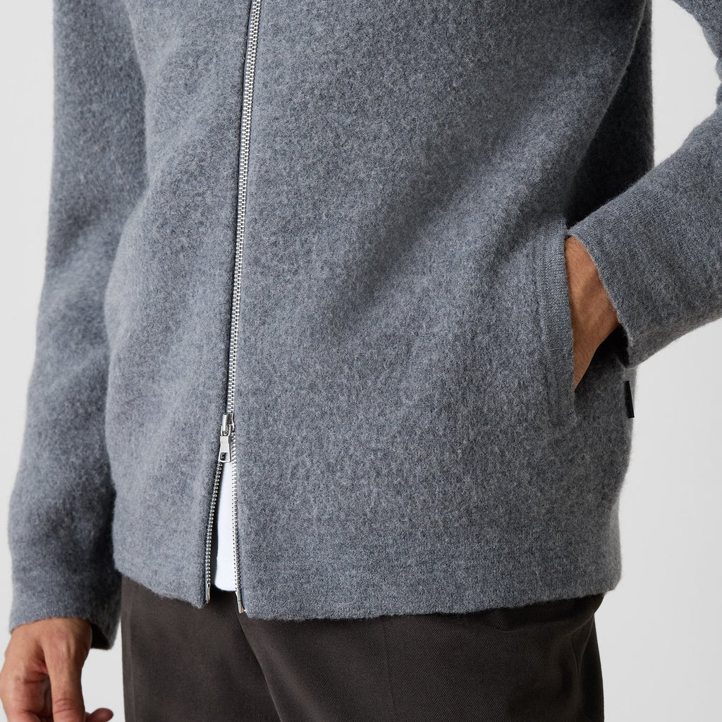 A close-up of a person wearing a heather gray zip-up jacket with side pockets, paired with dark trousers, against a light gray background. Oliver Sweeney Cardigan available at Sakks Menswear in Southampton.