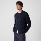 A man wearing a dark navy blue knit button-up cardigan over a white undershirt, paired with navy trousers, photographed against a light gray studio background. Oliver Sweeney cardigan available at Sakks Menswear in Southampton.