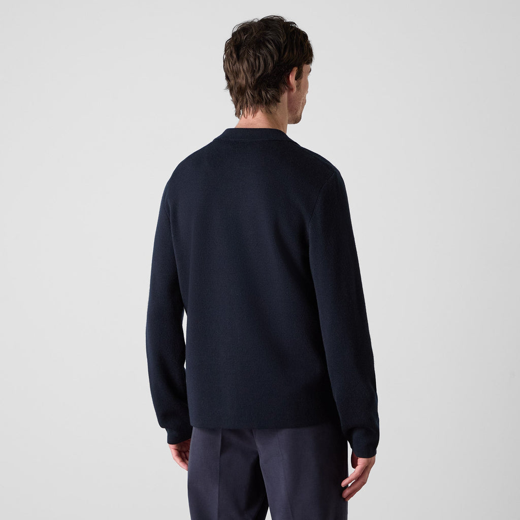 A man viewed from the back wearing a dark navy long-sleeve knit sweater and matching navy trousers against a light gray studio background. Oliver Sweeney cardigan available at Sakks Menswear in Southampton.