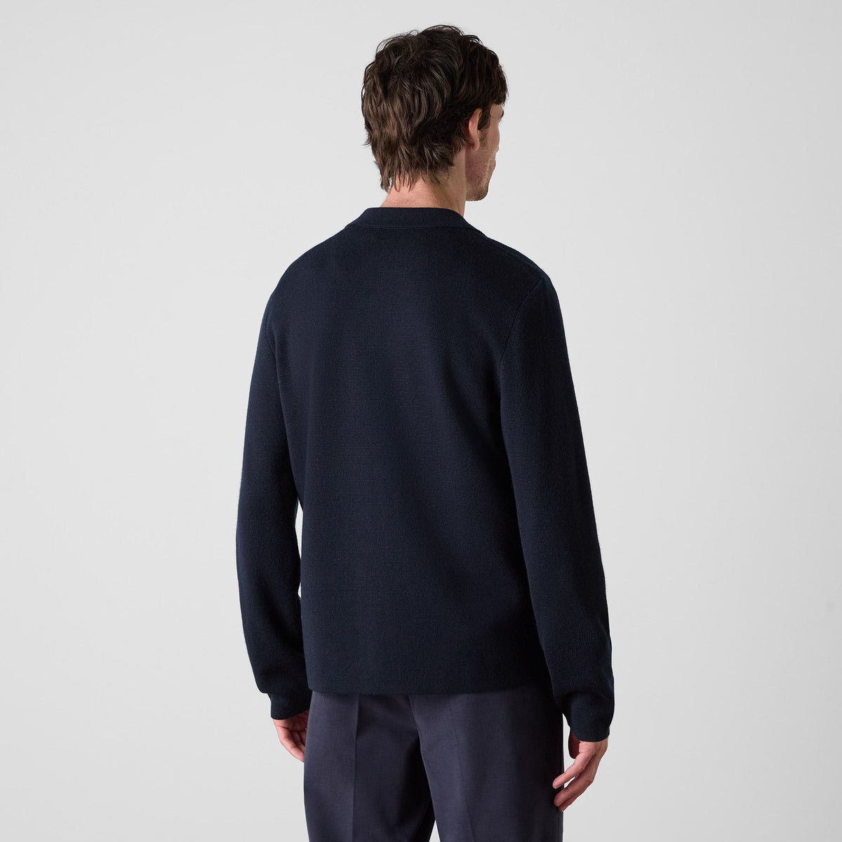 A man viewed from the back wearing a dark navy long-sleeve knit sweater and matching navy trousers against a light gray studio background. Oliver Sweeney cardigan available at Sakks Menswear in Southampton.