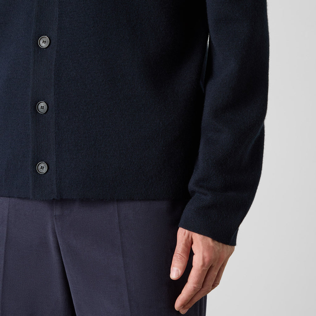 Close-up of a person wearing a dark navy knit button-front cardigan with dark buttons and navy trousers, shown against a light gray studio background. Oliver Sweeney cardigan available at Sakks Menswear in Southampton.