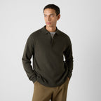 A man wearing a dark olive green long-sleeve polo knit sweater over a gray t-shirt and brown pants, standing against a light gray studio background. Oliver Sweeney Knitwear available at Sakks Menswear in Southampton.