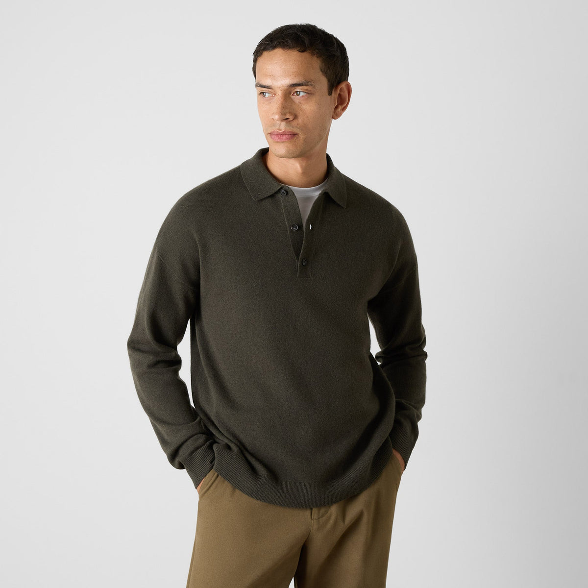 A man wearing a dark olive green long-sleeve polo knit sweater over a gray t-shirt and brown pants, standing against a light gray studio background. Oliver Sweeney Knitwear available at Sakks Menswear in Southampton.