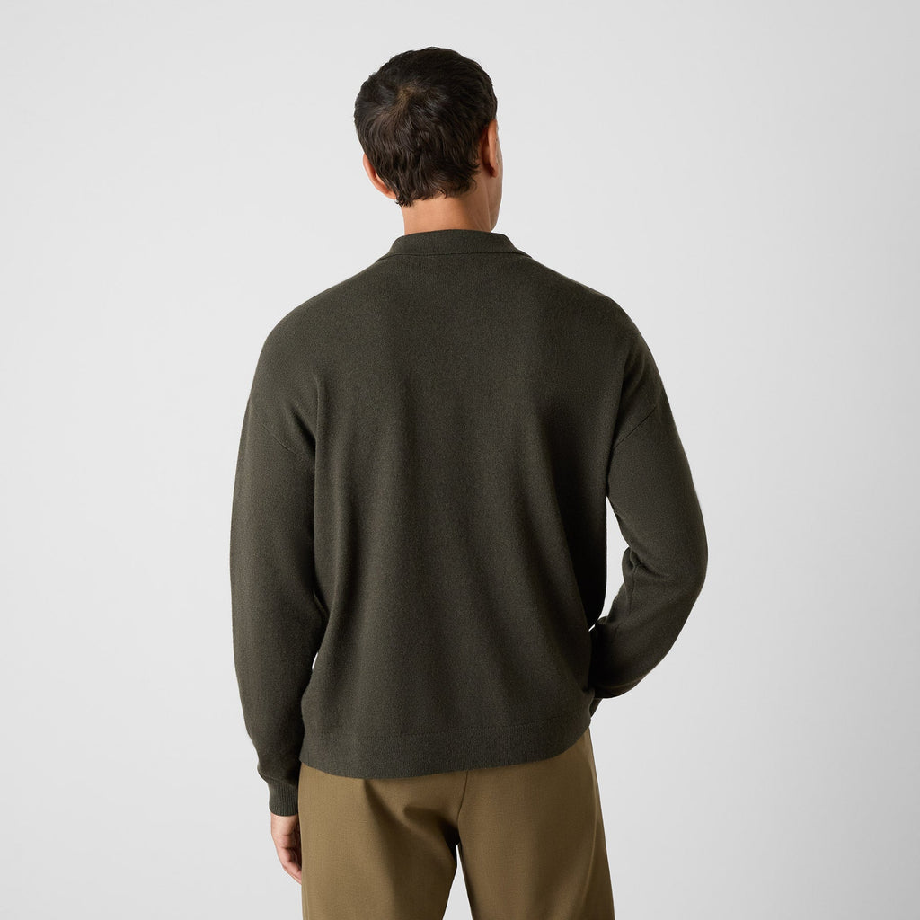 A man viewed from behind wearing a dark olive green knit sweater and brown trousers, standing against a light gray studio background. Oliver Sweeney Knitwear available at Sakks Menswear in Southampton.