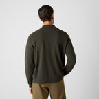 A man viewed from behind wearing a dark olive green knit sweater and brown trousers, standing against a light gray studio background. Oliver Sweeney Knitwear available at Sakks Menswear in Southampton.