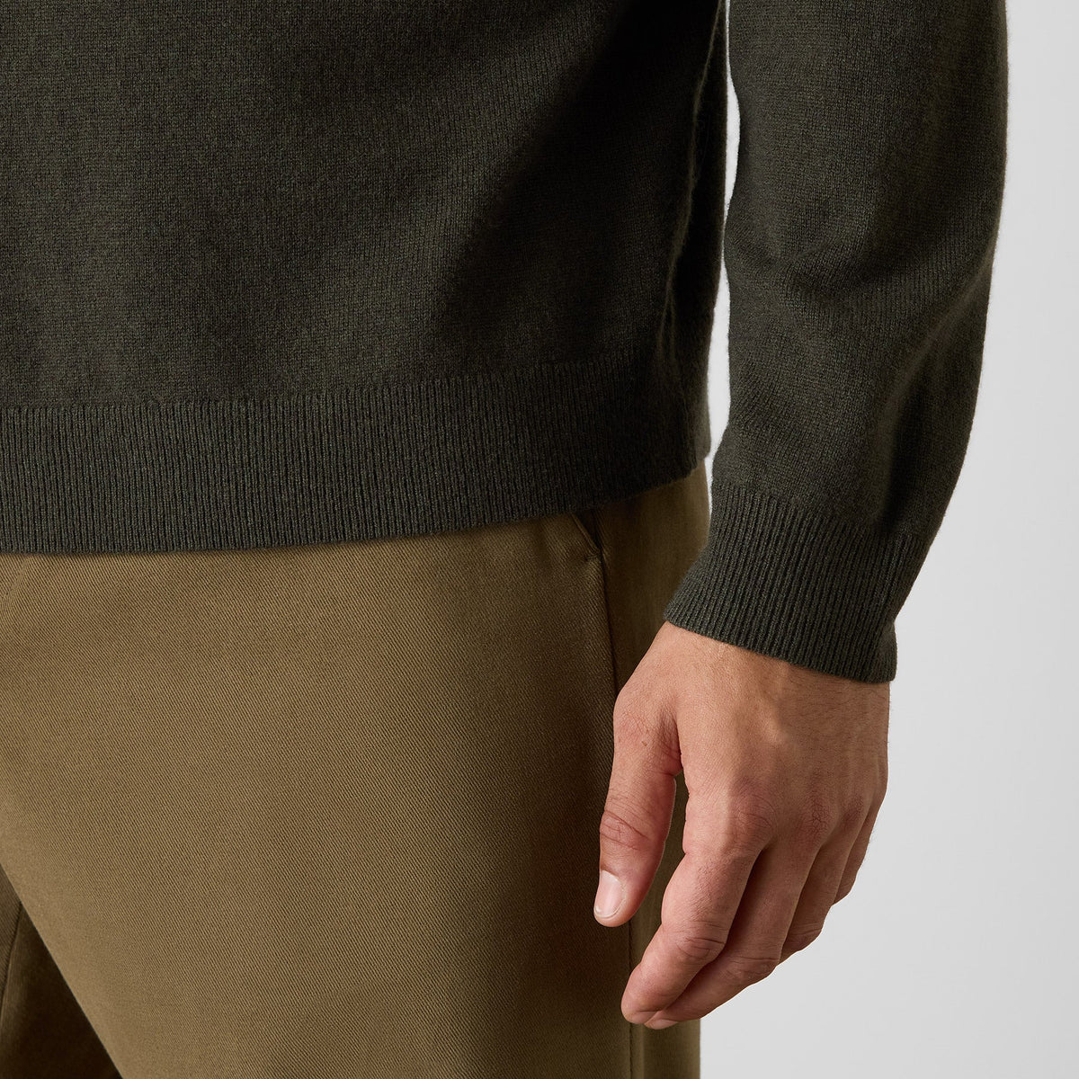 Close-up of a person wearing a dark olive green knit sweater with ribbed cuffs and hem, paired with tan trousers, against a light gray background. Oliver Sweeney Knitwear available at Sakks Menswear in Southampton.
