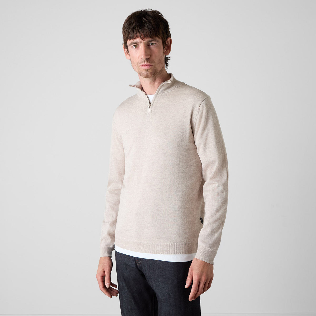 A man wearing a light beige quarter-zip pullover sweater and dark trousers, standing against a plain light gray studio background. Oliver Sweeney knitwear available at Sakks Menswear in Southampton.