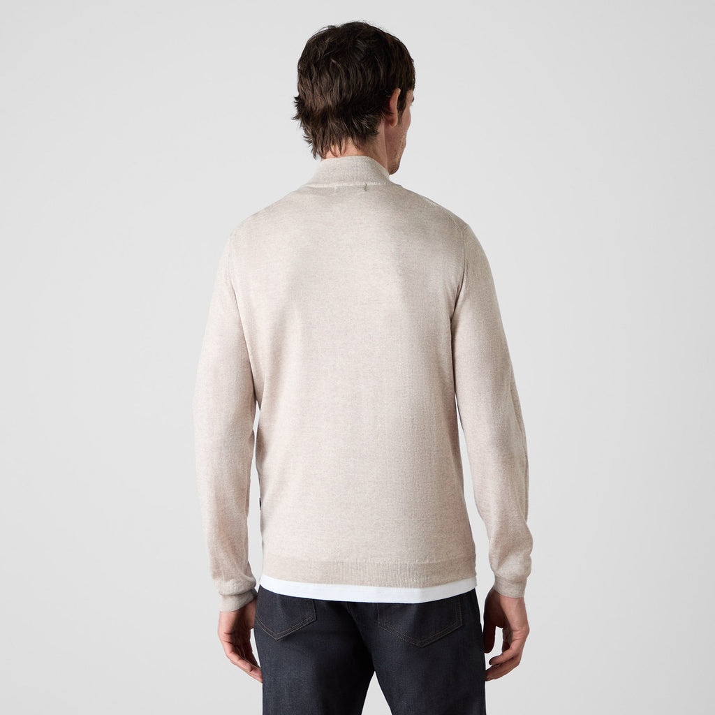 A man viewed from behind wearing a light beige knit sweater over a white undershirt and dark jeans, set against a plain light gray studio background. Oliver Sweeney knitwear available at Sakks Menswear in Southampton.