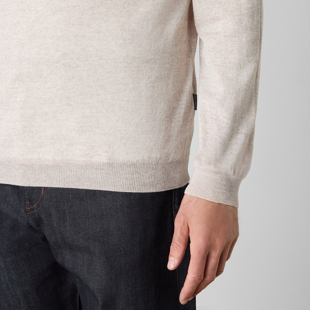 Close-up of a person wearing a light beige knit sweater with dark blue jeans, showing the ribbed hem and cuffs, and the person's hand. Oliver Sweeney knitwear available at Sakks Menswear in Southampton.