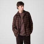 A man wearing a brown and dark brown plaid overshirt over a matching brown t-shirt and dark brown pants, standing against a light gray studio background. Oliver Sweeney Shirt Jacket available at Sakks Menswear in Southampton.