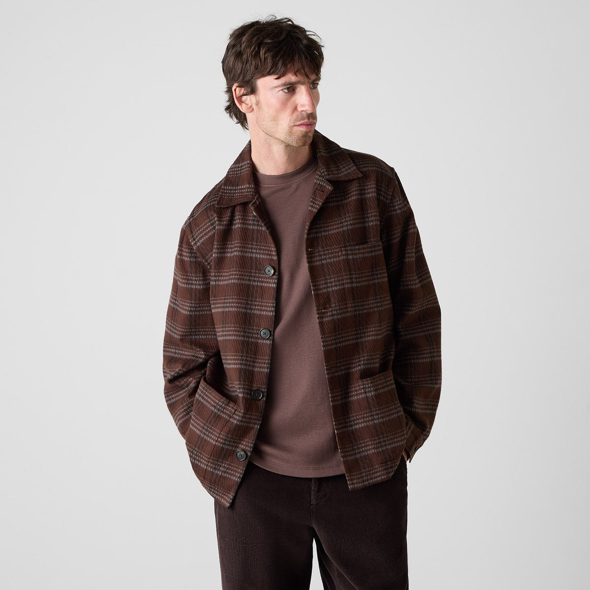 A man wearing a brown and dark brown plaid overshirt over a matching brown t-shirt and dark brown pants, standing against a light gray studio background. Oliver Sweeney Shirt Jacket available at Sakks Menswear in Southampton.