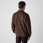 A man viewed from behind wearing a brown and dark gray plaid flannel shirt jacket, paired with dark brown trousers, against a light gray studio background. Oliver Sweeney Shirt Jacket available at Sakks Menswear in Southampton.