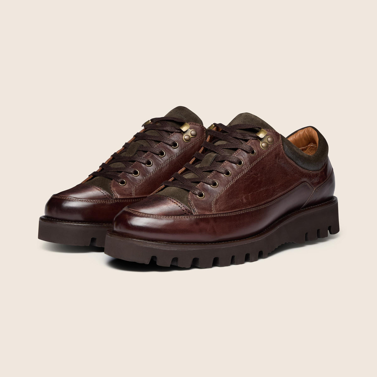 A pair of dark brown leather low-top sneakers with chunky lug soles, dark laces, and brass eyelets, displayed on a light beige background. Oliver Sweeney Boots available at Sakks Menswear in Southampton.
