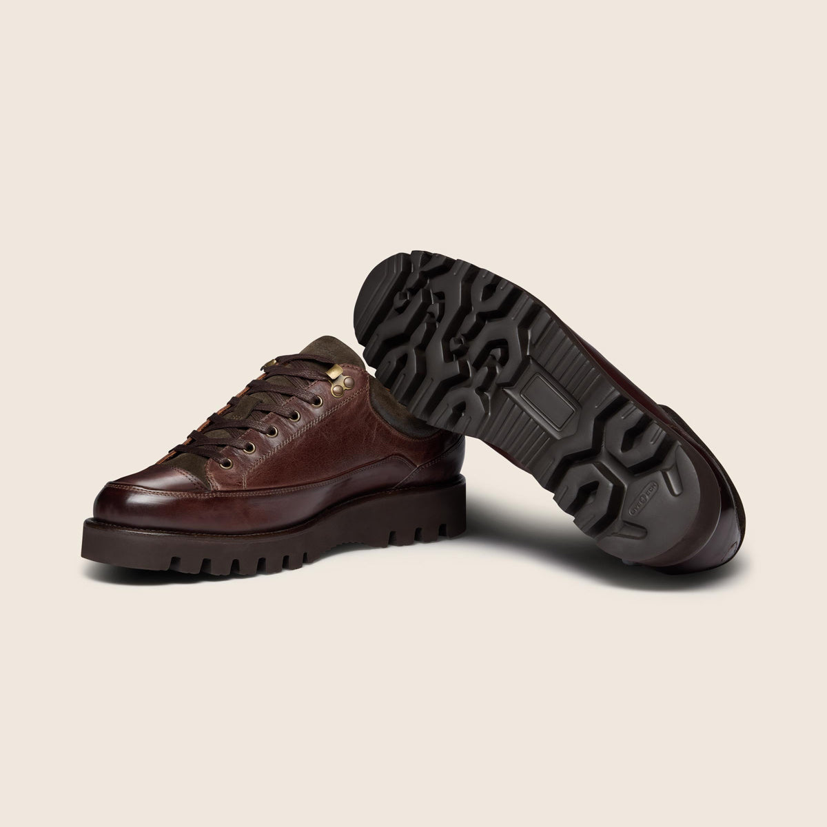 A pair of dark brown leather low-top lace-up shoes with chunky lug soles, shown on a light beige background. One shoe is standing upright while the other is tilted on its side to display the textured black sole. Oliver Sweeney Boots available at Sakks Menswear in Southampton.