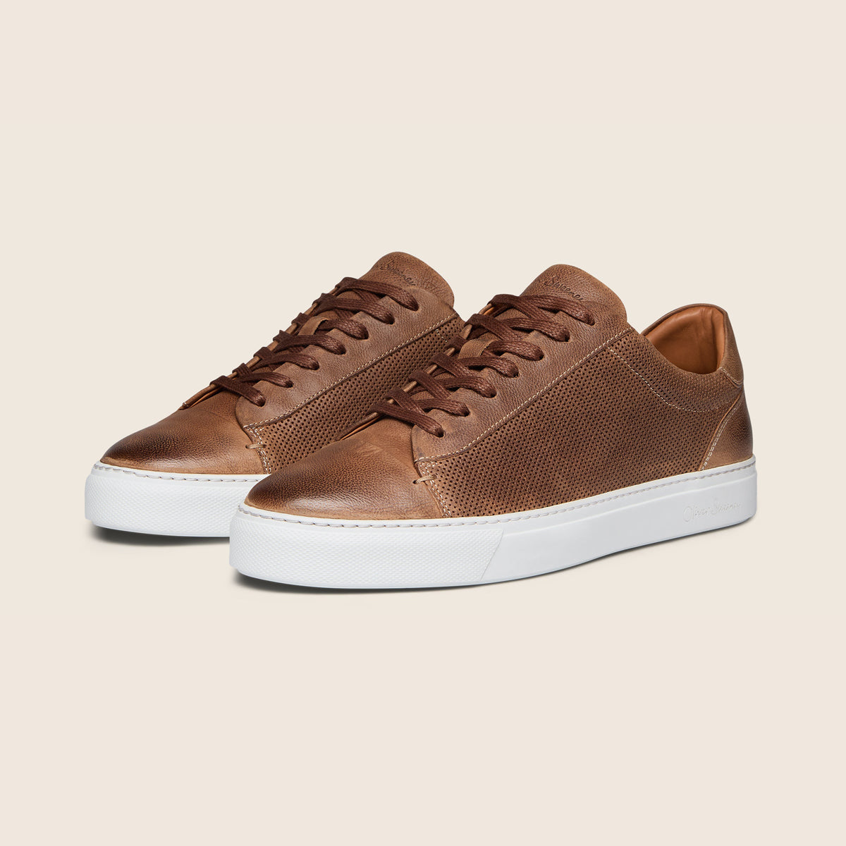 A pair of low-top brown leather sneakers with textured perforated panels, matching brown laces, and thick white rubber soles, displayed on a light beige background. Oliver Sweeney Trainers available at Sakks Menswear in Southampton.