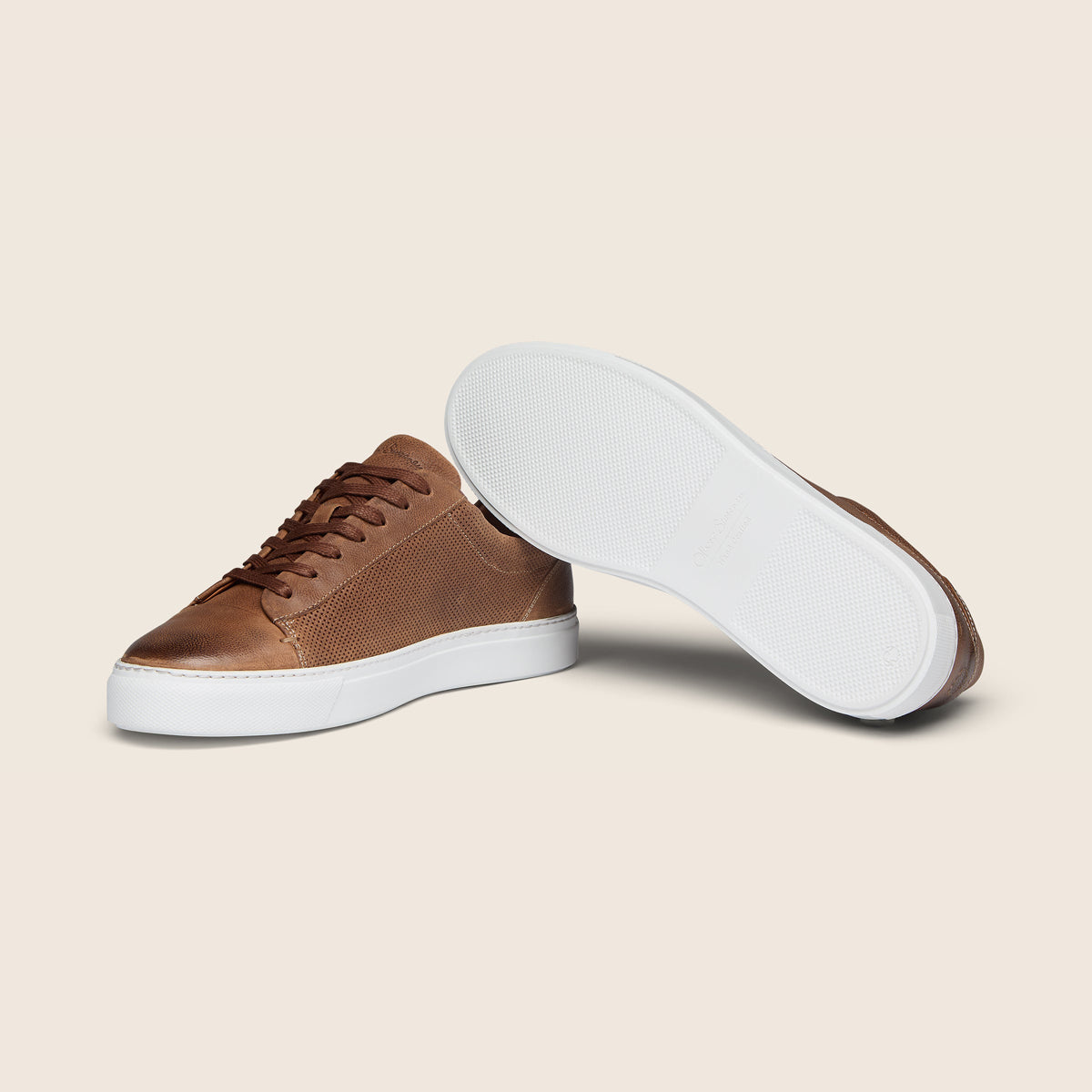 A pair of low-top brown leather sneakers, with one shoe standing upright in side view and the other lying on its side to show the white outsole, set against a solid beige background. Oliver Sweeney Trainers available at Sakks Menswear in Southampton.
