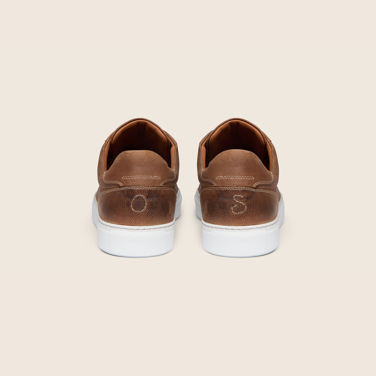 A pair of brown leather low-top sneakers shown from the back, featuring white rubber soles and a debossed 'OS' logo on the heel, set against a solid light beige background. Oliver Sweeney Trainers available at Sakks Menswear in Southampton.