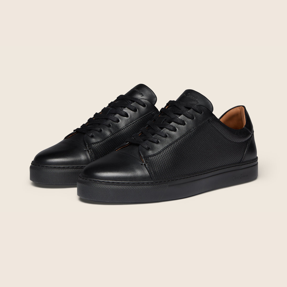 A pair of black leather low-top sneakers with textured perforated panels on the sides, black laces, and thick black rubber soles, displayed on a light beige background. Oliver Sweeney Trainers available at Sakks Menswear in Southampton.