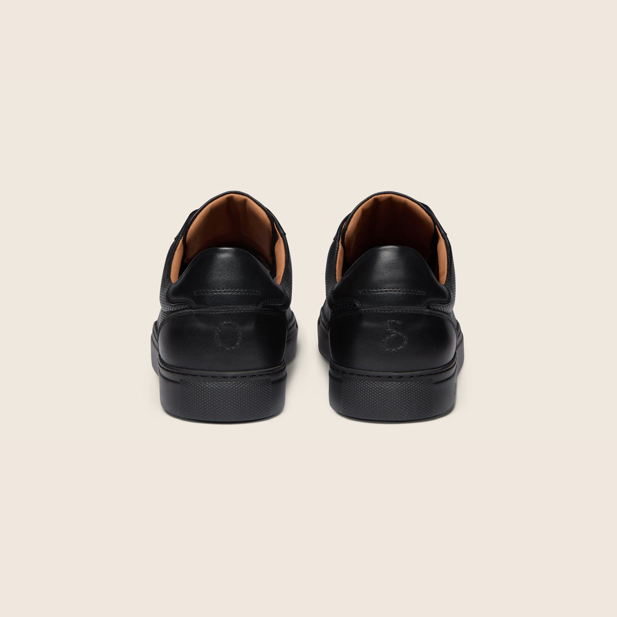 A pair of black leather low-top sneakers shown from the back, featuring a black rubber sole and a contrasting tan interior lining, set against a solid light beige background. Oliver Sweeney Trainers available at Sakks Menswear in Southampton.