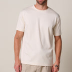 a model stands in front of a light grey background. He wears a raw ehite pima cotton t-shirt matched with beige linen trousers. Profuomo T-Shirts are available at Sakks Menswear in Southampton.