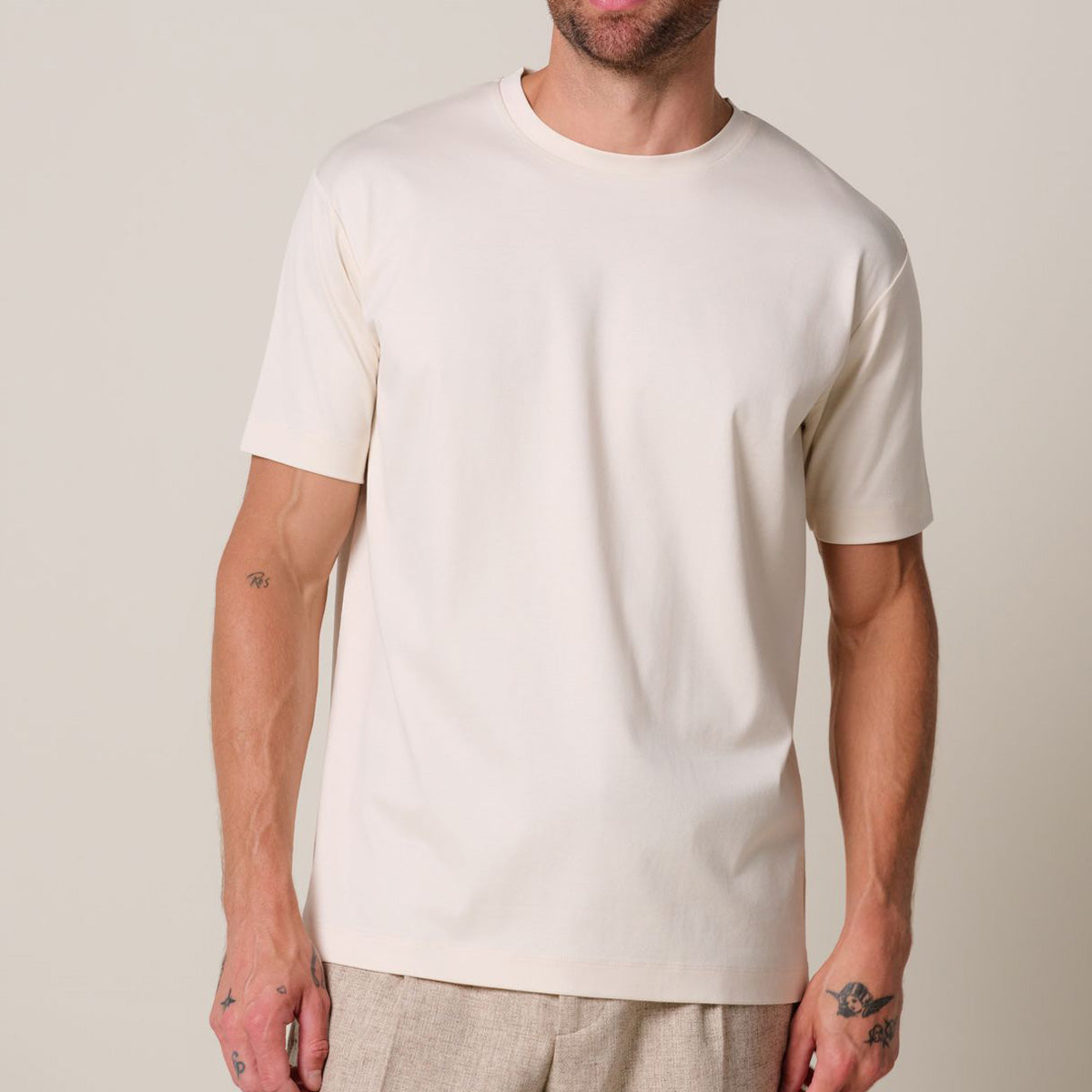 a model stands in front of a light grey background. He wears a raw ehite pima cotton t-shirt matched with beige linen trousers. Profuomo T-Shirts are available at Sakks Menswear in Southampton.