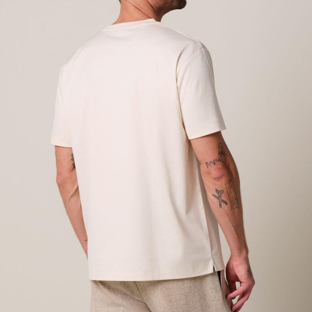 a model stands in front of a light grey background. He wears a raw ehite pima cotton t-shirt matched with beige linen trousers. Profuomo T-Shirts are available at Sakks Menswear in Southampton.