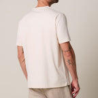 a model stands in front of a light grey background. He wears a raw ehite pima cotton t-shirt matched with beige linen trousers. Profuomo T-Shirts are available at Sakks Menswear in Southampton.