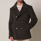 A man wearing a dark brown double-breasted peacoat over a cream shirt, photographed against a plain light beige studio background. Profuomo coat available at Sakks Menswear in Southampton.