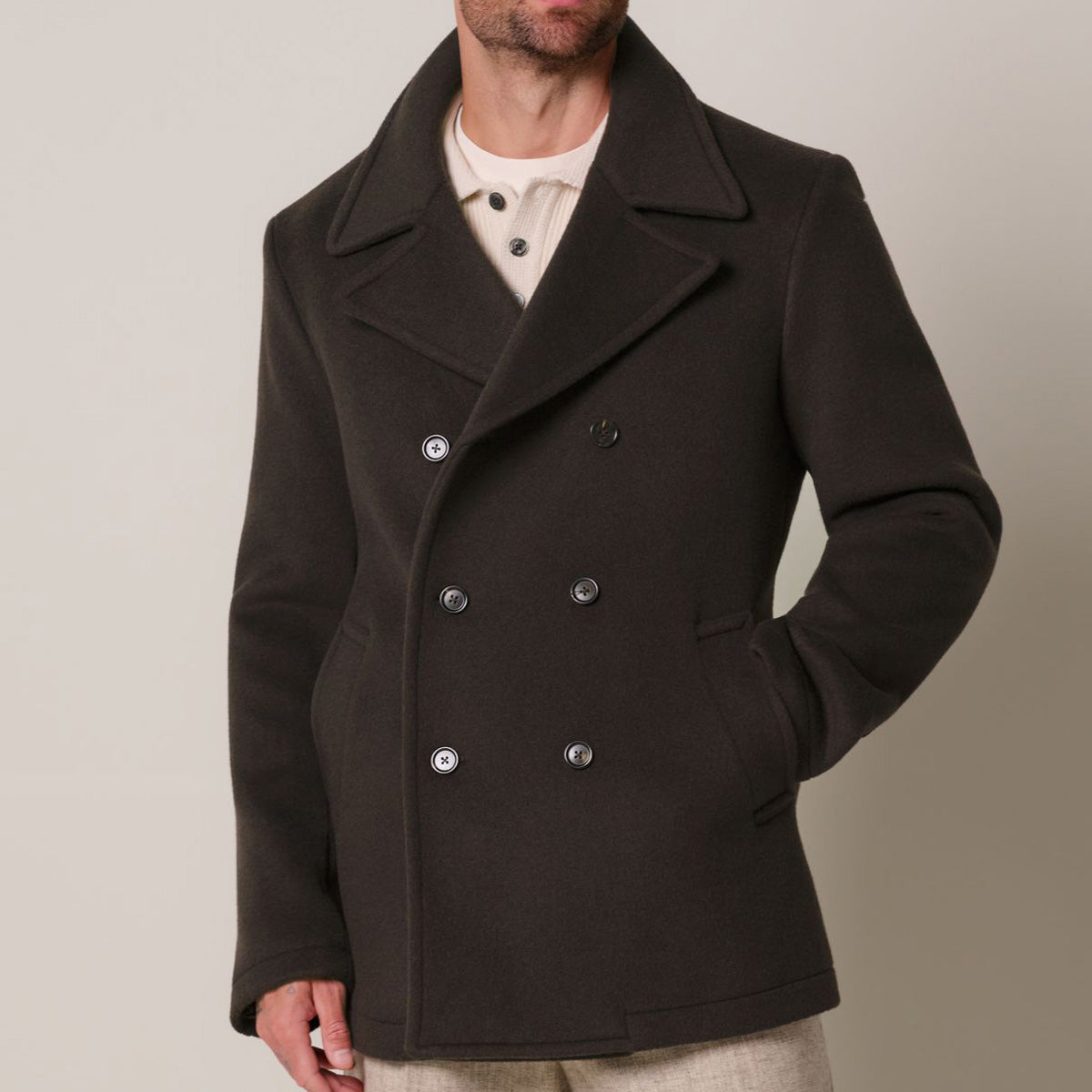 A man wearing a dark brown double-breasted peacoat over a cream shirt, photographed against a plain light beige studio background. Profuomo coat available at Sakks Menswear in Southampton.