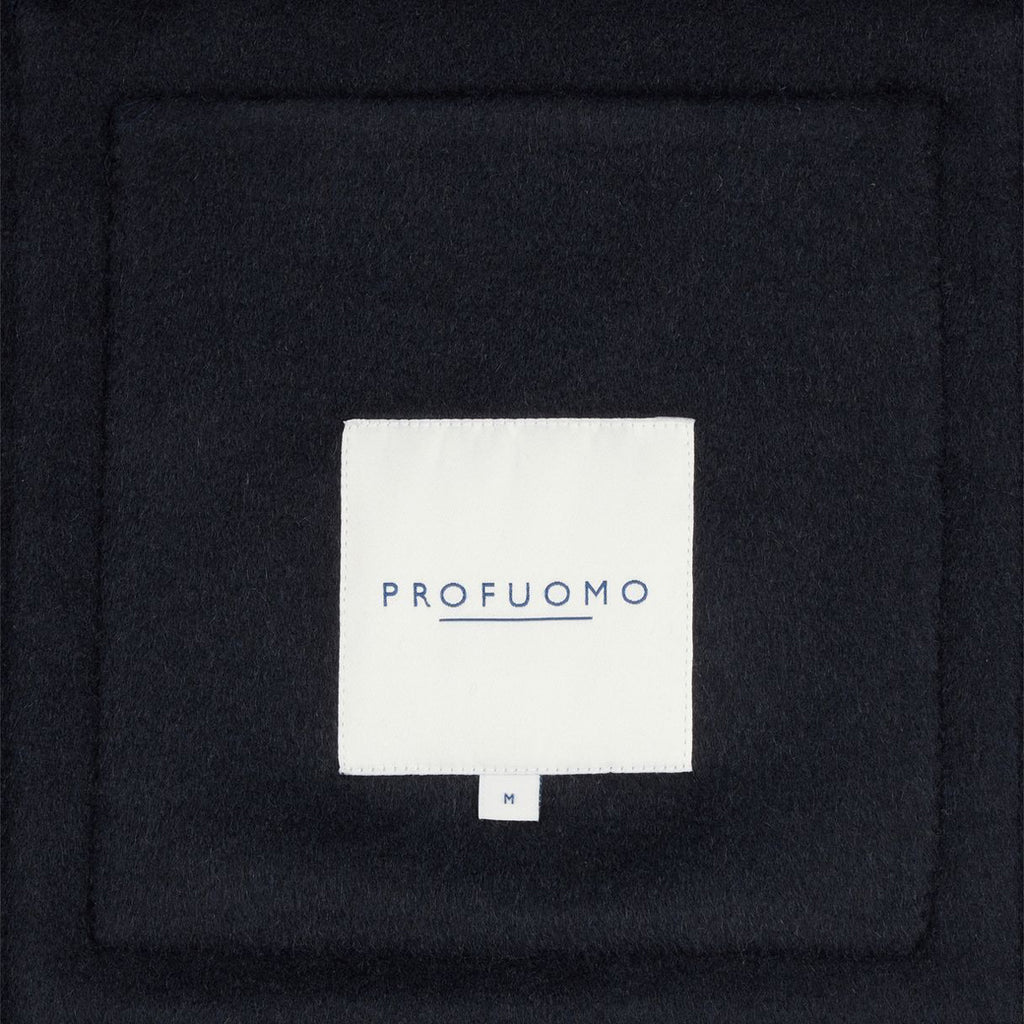 A close-up of a dark navy or black textured fabric with a centered white square label reading 'PROFUOMO' in blue text.  Profuomo Coat available at Sakks Menswear in Southampton.
