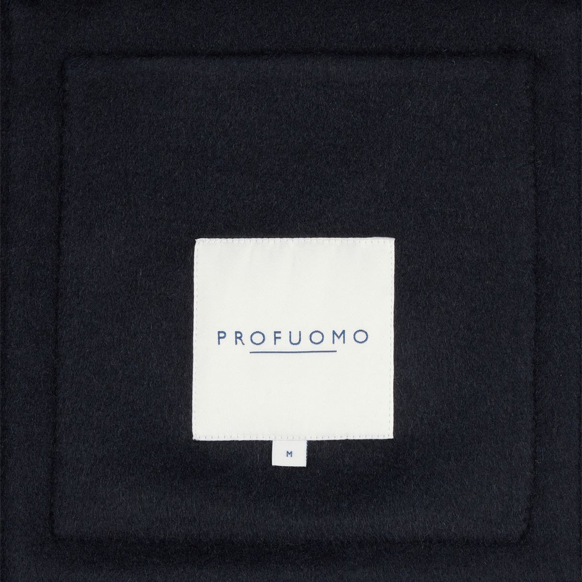 A close-up of a dark navy or black textured fabric with a centered white square label reading 'PROFUOMO' in blue text.  Profuomo Coat available at Sakks Menswear in Southampton.