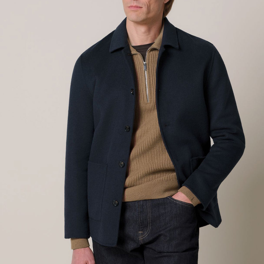 A man wearing a dark navy blue jacket over a tan ribbed quarter-zip sweater and dark jeans, photographed against a plain light beige studio background.  Profuomo Coat available at Sakks Menswear in Southampton.