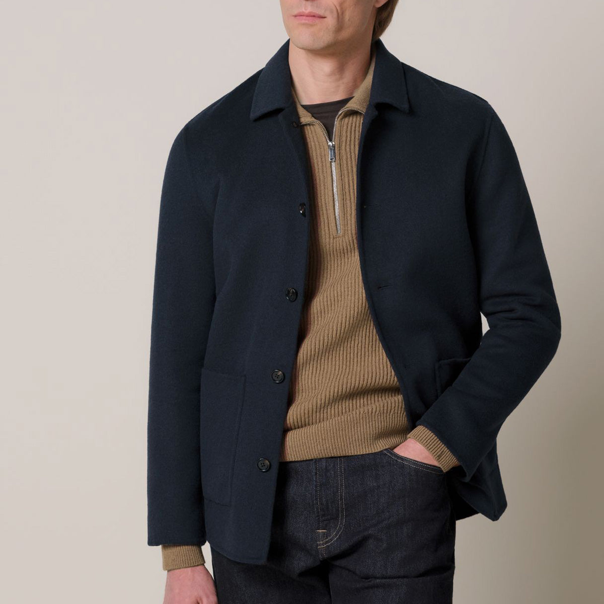 A man wearing a dark navy blue jacket over a tan ribbed quarter-zip sweater and dark jeans, photographed against a plain light beige studio background.  Profuomo Coat available at Sakks Menswear in Southampton.