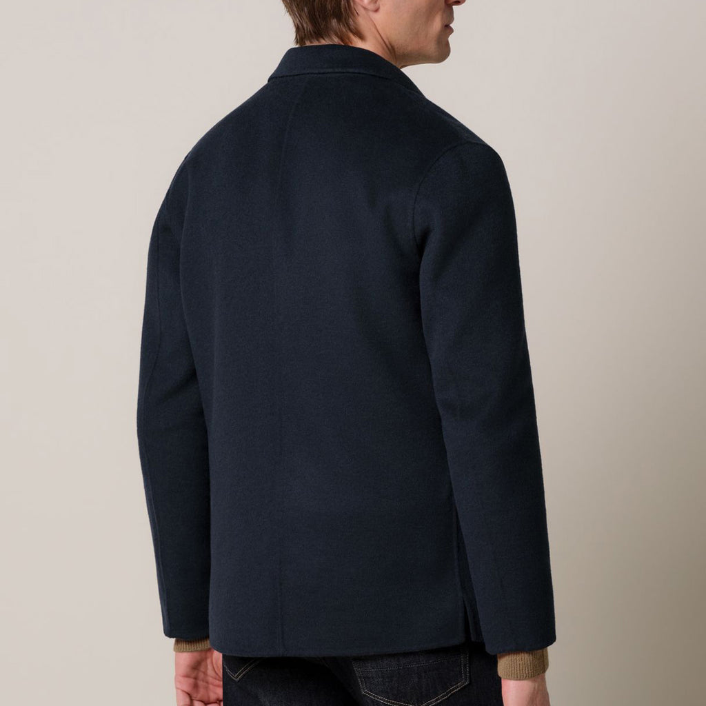 A man shown from the back wearing a dark navy blue jacket, photographed against a plain light beige studio background. Profuomo Coat available at Sakks Menswear in Southampton.