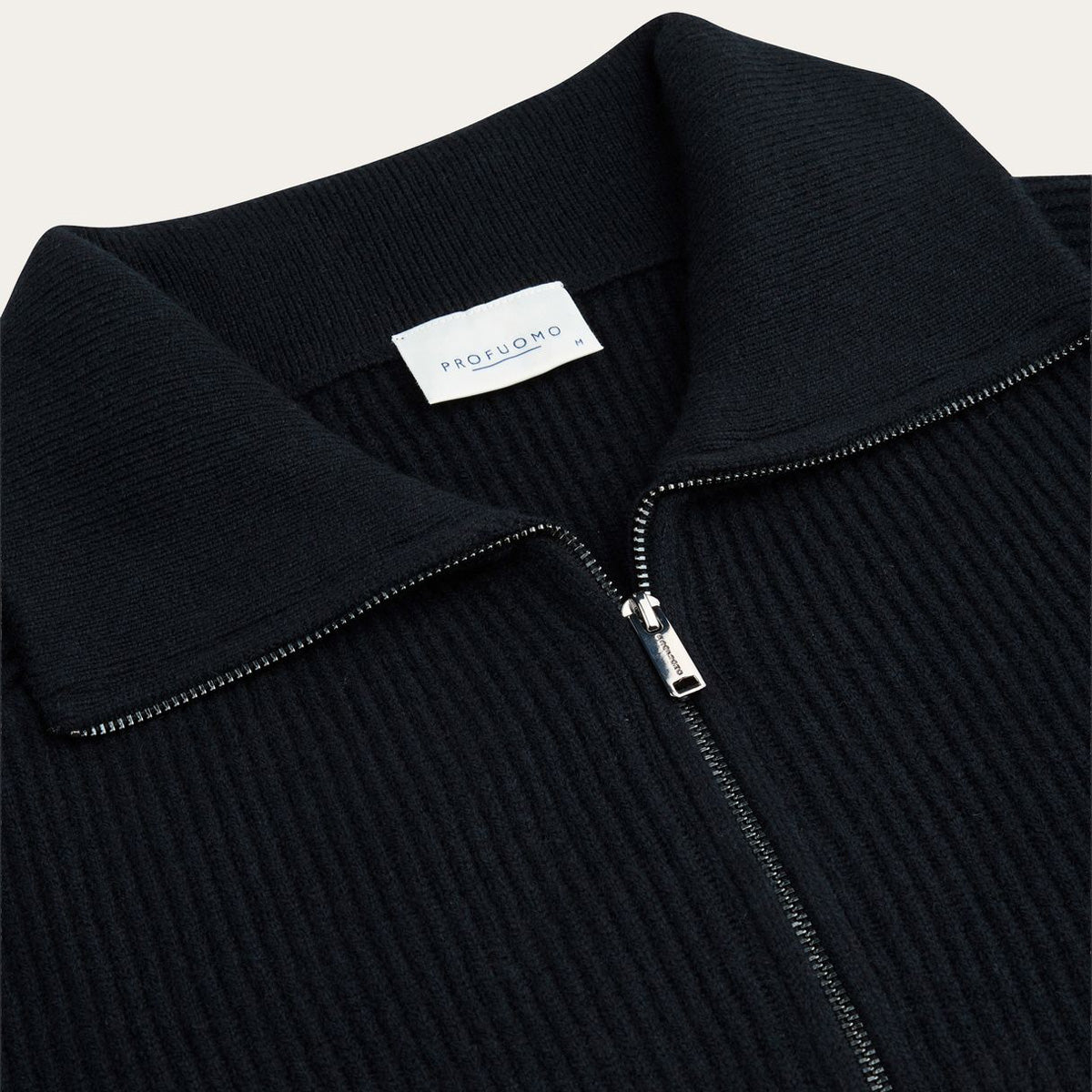 Close-up of a ribbed knit zip-up sweater collar in black, with a white neck label reading PROFUOMO and a silver zipper pull. Profuomo Knitwear available at Sakks Menswear in Southampton.