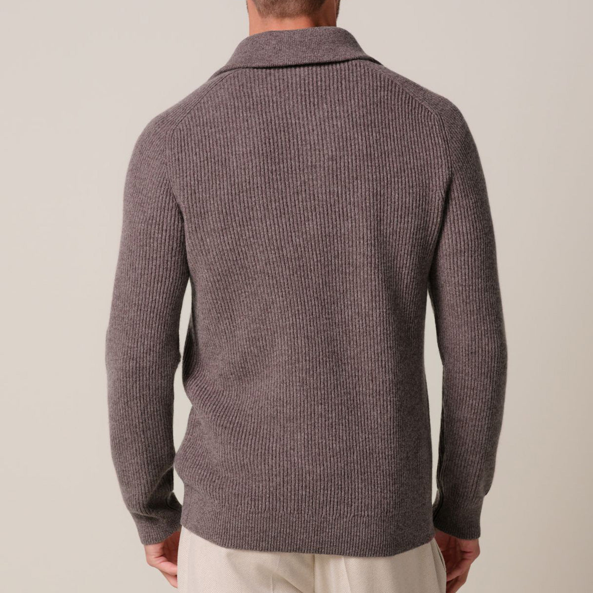 A man viewed from the back wearing a ribbed knit polo-style sweater in a muted taupe-brown color, paired with light beige trousers, against a plain light beige studio background. Profuomo Knitwear available at Sakks Menswear in Southampton.