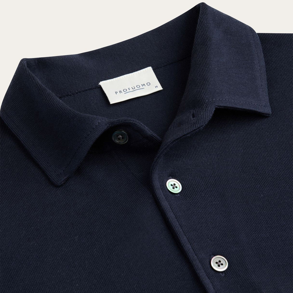 Close-up of a dark navy polo shirt collar with a white PROFUOMO brand label inside the neck, featuring silver buttons on a light beige background. Profuomo Polos available at Sakks Menswear in Southampton.