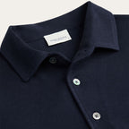 Close-up of a dark navy polo shirt collar with a white PROFUOMO brand label inside the neck, featuring silver buttons on a light beige background. Profuomo Polos available at Sakks Menswear in Southampton.