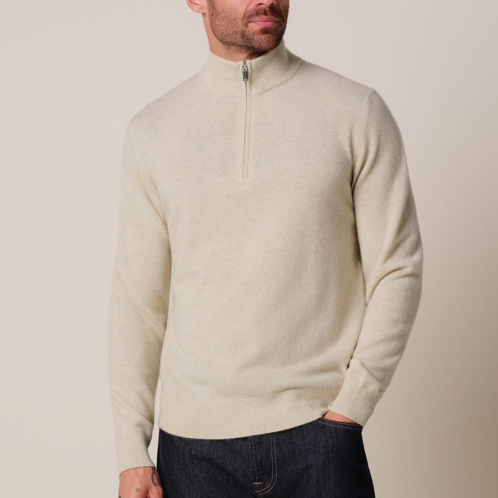 A man wearing a light beige half-zip knit sweater with dark blue jeans, photographed against a solid light beige background. Profuomo knitwear available at Sakks Menswear in Southampton.
