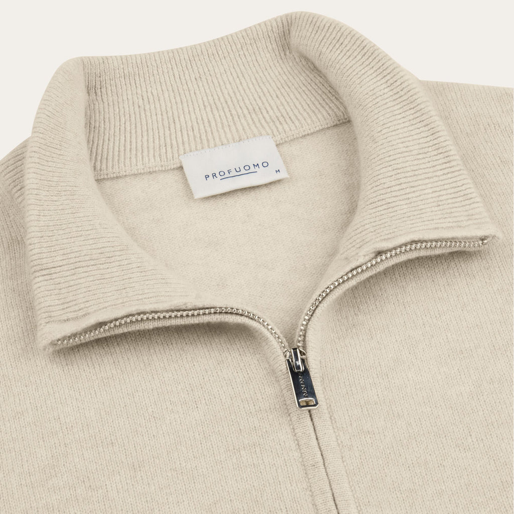 Close-up of a ribbed knit quarter-zip sweater collar in a light warm beige/cream tone, with a visible PROFUOMO neck label and a silver zipper pull. Profuomo knitwear available at Sakks Menswear in Southampton.