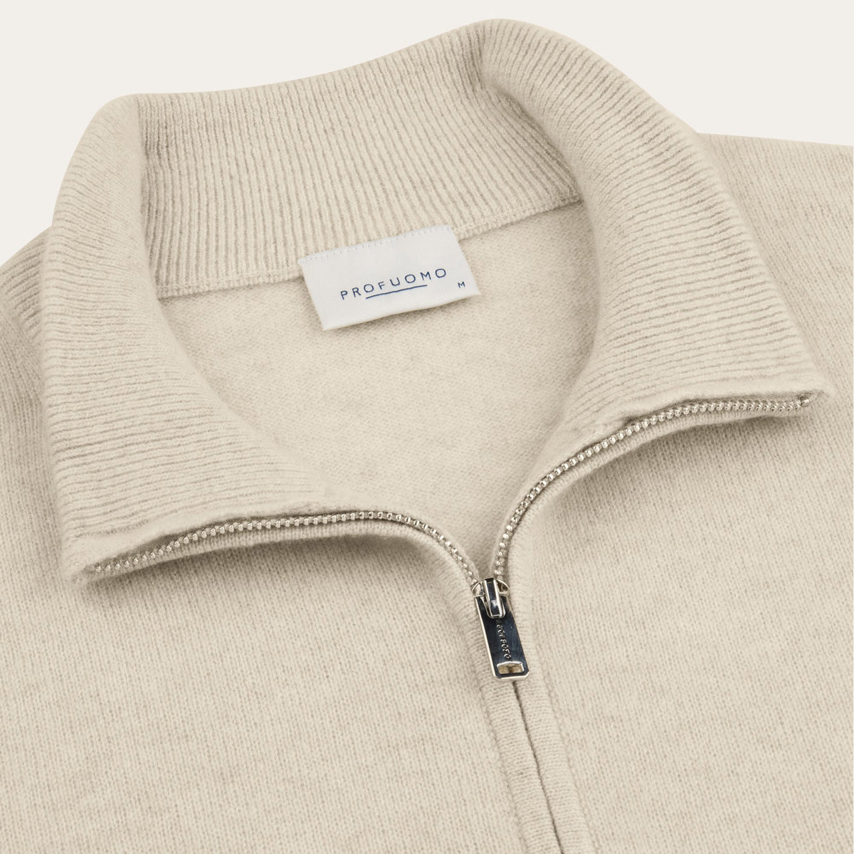 Close-up of a ribbed knit quarter-zip sweater collar in a light warm beige/cream tone, with a visible PROFUOMO neck label and a silver zipper pull. Profuomo knitwear available at Sakks Menswear in Southampton.