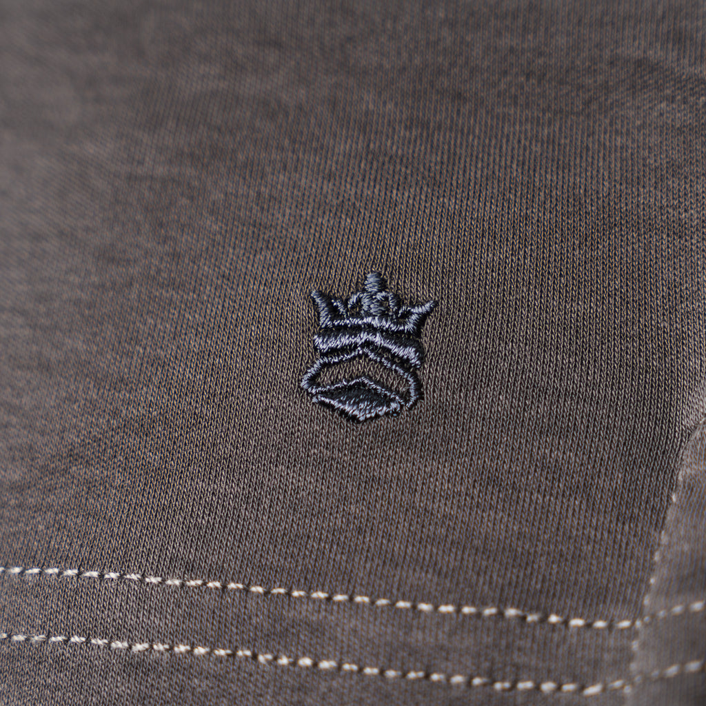 Close-up of a dark gray textured fabric with a small embroidered crown logo in black and gray, plus white contrast stitching near the bottom. Thomas Maine T-Shirts available at Sakks Menswear in Southampton.