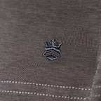 Close-up of a dark gray textured fabric with a small embroidered crown logo in black and gray, plus white contrast stitching near the bottom. Thomas Maine T-Shirts available at Sakks Menswear in Southampton.